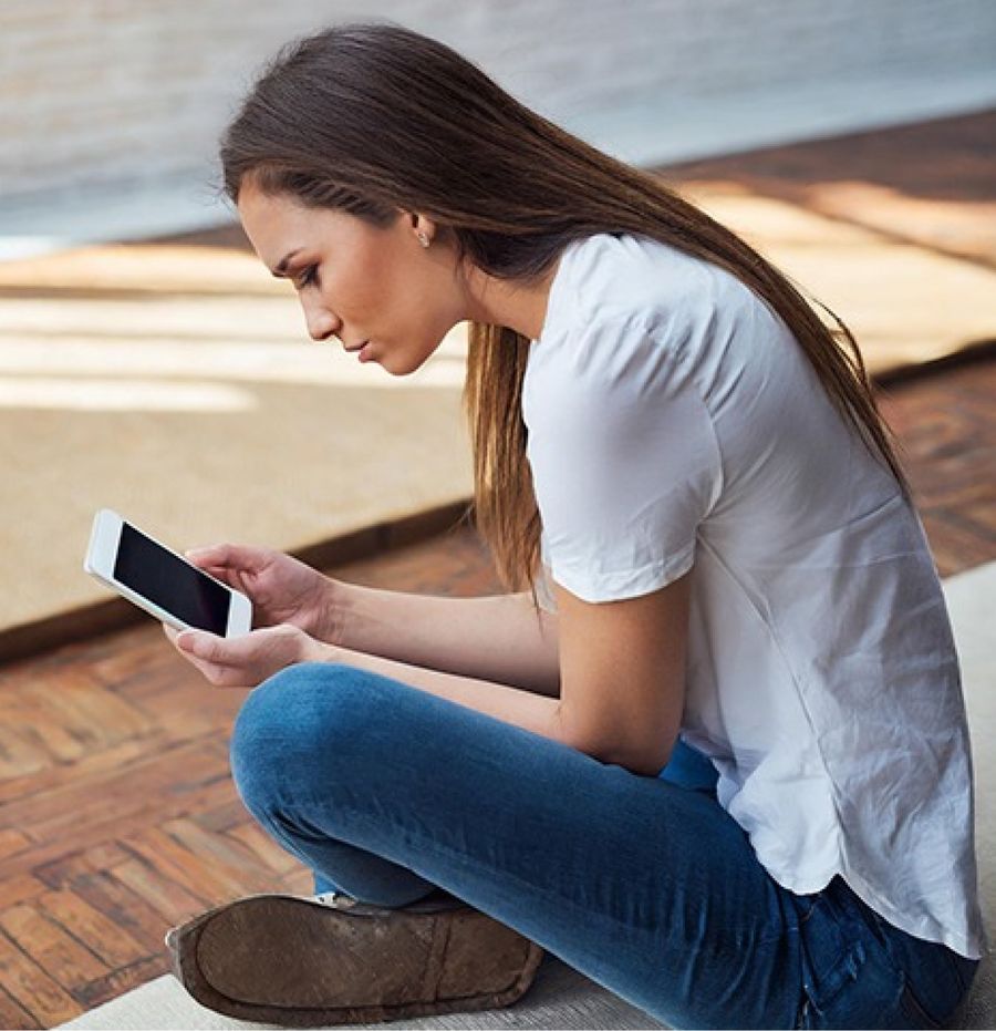 A young woman sitting cross legged on the floor, leaning over her phone