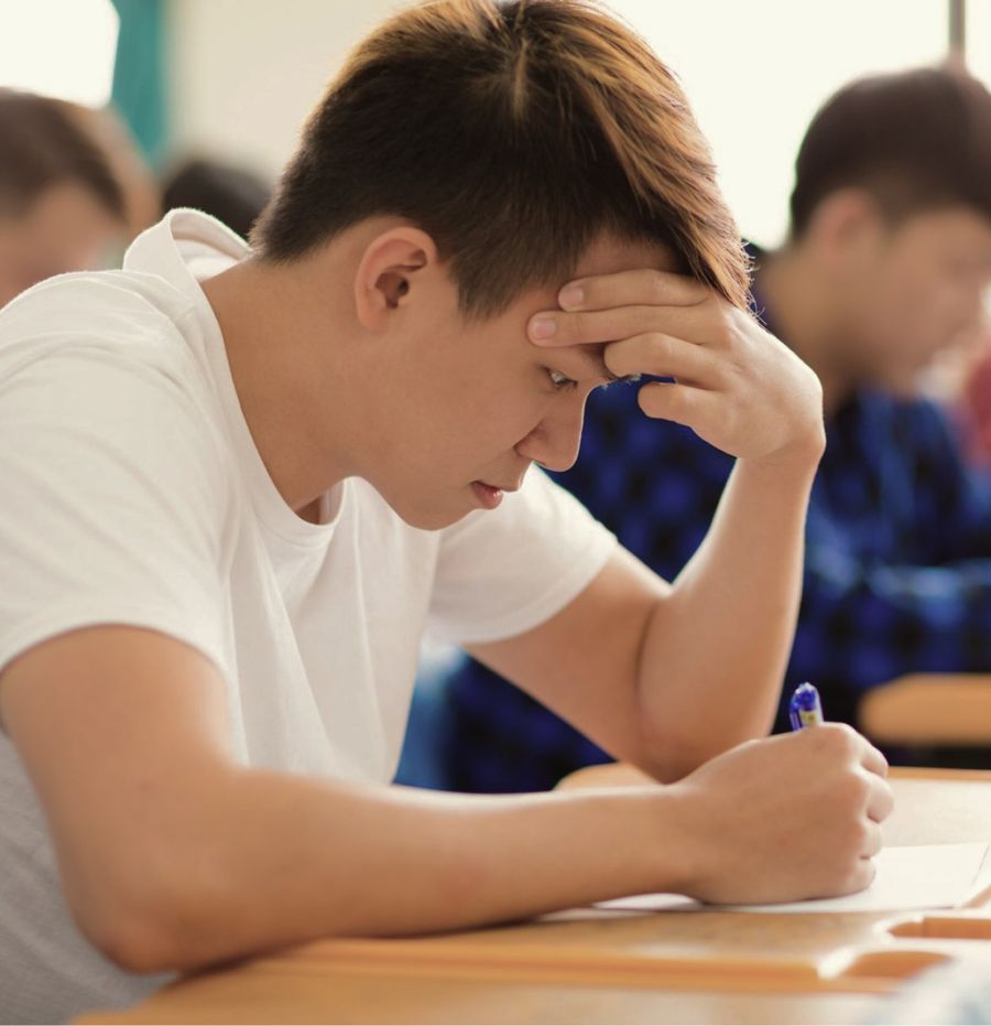 A student writing in a classroom, head bent low over the desk